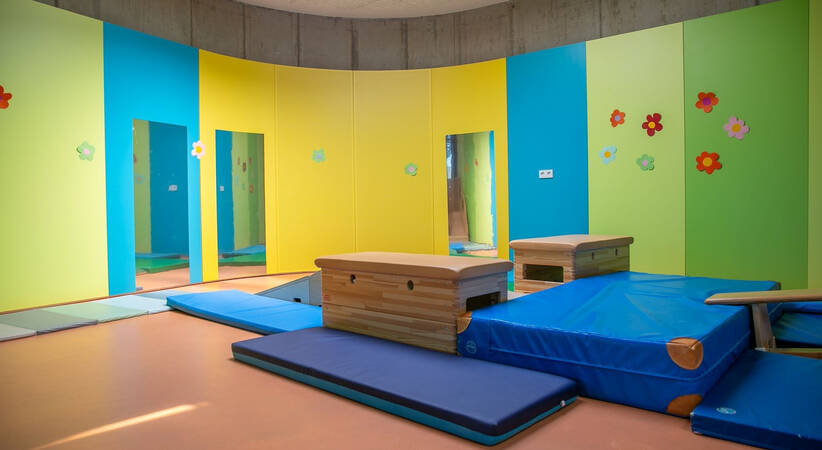 Der Raum zeigt eine bunte Sporthalle mit Matten am Boden, bunten Möbeln und Wänden, die mit Blumen dekoriert sind.