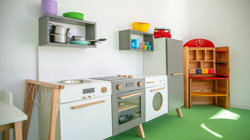 Ein heller Raum mit einer Spielzeugküche aus Holz, bestehend aus Herd, Backofen und Waschmaschine, sowie einem Regal mit bunten Elementen und einer Spieluhr auf grünem Teppichboden.