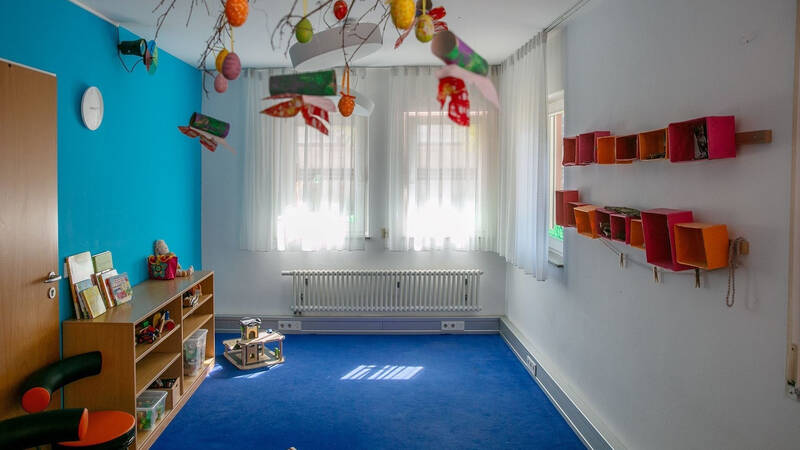 Ein heller Raum mit blauen Wänden und Teppich, in dem bunte Dekorationen von der Decke hängen, ein Regal voller Spielzeug steht und an der Wand bunte Kästen angebracht sind.