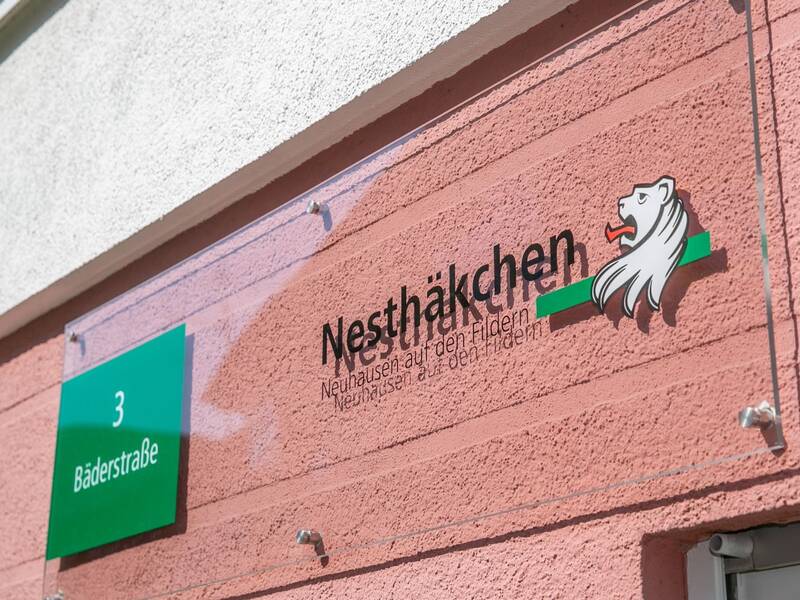 Ein Schild an einer rosa Wand zeigt den Straßennamen "Bäderstraße 3" und das Logo einer Einrichtung namens "Nesthäkchen" mit einem stilisierten Löwenkopf.