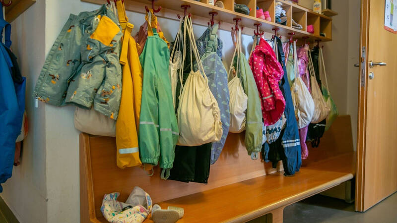 In einem Eingangsbereich einer Kindertagesstätte hängen bunte Jacken und Taschen über einer Holzbank an Haken.