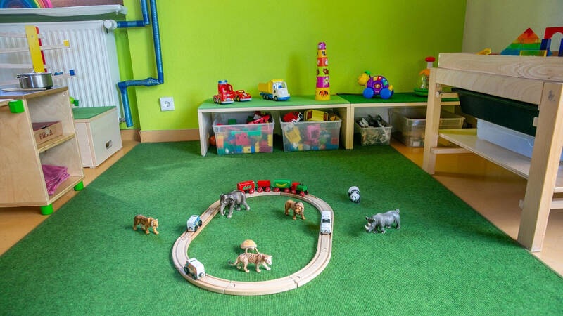 Ein Spielzimmer mit grünem Teppich, auf dem eine Holzeisenbahn kreisförmig aufgebaut ist und von Tierfiguren umgeben wird, während im Hintergrund Regale mit Spielzeug und Kisten sichtbar sind.