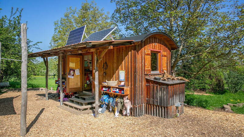 Ein kleines, hölzernes Häuschen mit Solarpanelen auf dem Dach steht umgeben von Bäumen und Wiesen.
