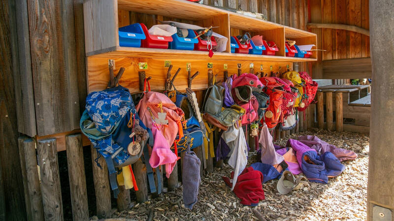Mehrere bunte Kinderrucksäcke und Jacken hängen ordentlich an Haken unter einem Regal mit farbigen Schachteln in einem Holzgebäude.