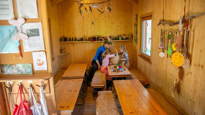 In einem holzverkleideten Raum sitzen drei Personen an einem Tisch, umgeben von dekorativen Bastelmaterialien und Holzmöbeln.