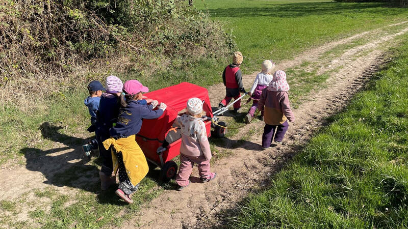 Eine Gruppe von Kindern zieht einen roten Bollerwagen über einen grasbewachsenen Weg neben einem Gebüsch.