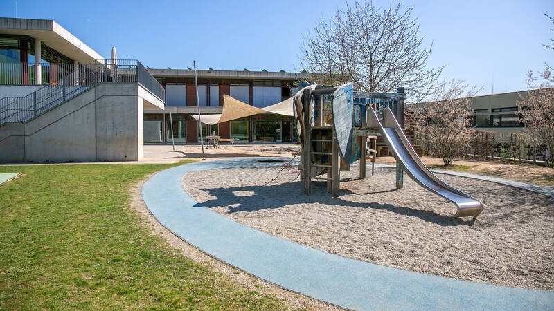 Der Spielplatz auf dem Bild zeigt eine Rutsche und Kletterelemente, umgeben von einem gepflasterten Weg und einer Wiese, mit einem modernen Gebäude im Hintergrund.