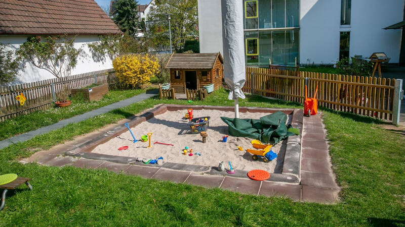 Ein Sandkasten mit verschiedenen bunten Spielzeugen, umgeben von einem Garten mit Gras und Zäunen, steht neben einem kleinen Holzhaus und einem geschlossenen Sonnenschirm.